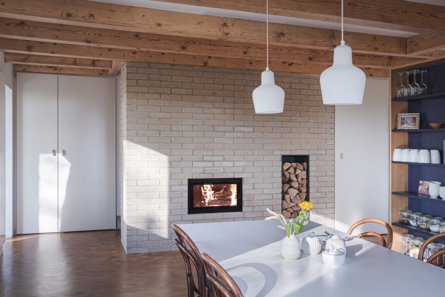 Séjour avec des solives en bois au plafond et un mur en brique crue à l’intérieur, avec un poêle à bois.