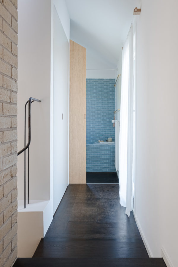 Couloir avec la salle de bains au fond et la rampe d’escalier en métal sur la gauche.
